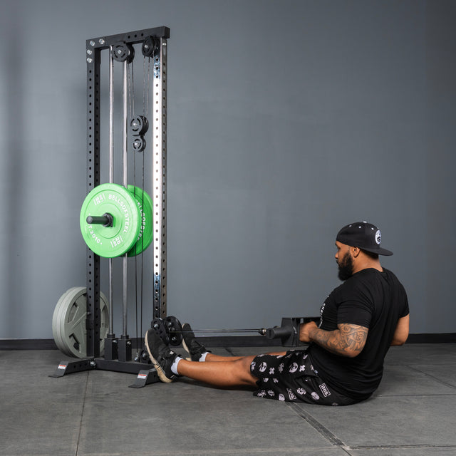 A man in a black shirt and shorts uses the Bells of Steel Arch Nemesis Tricep Bar with green weight plates in a gym to perform back exercises on a seated cable row machine.