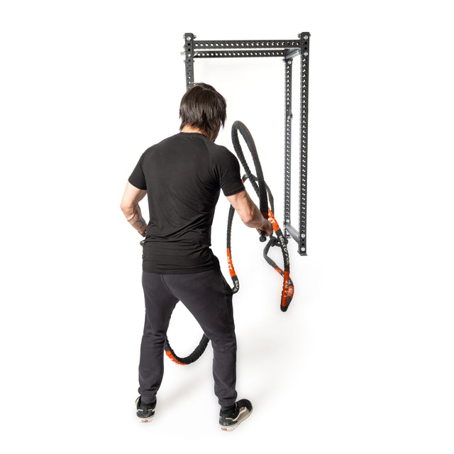 A person in black clothes uses a Battle Rope by Bells of Steel attached to a metal wall mount, showcasing a conditioning workout. The setup in the white space highlights the fitness equipment and posture.