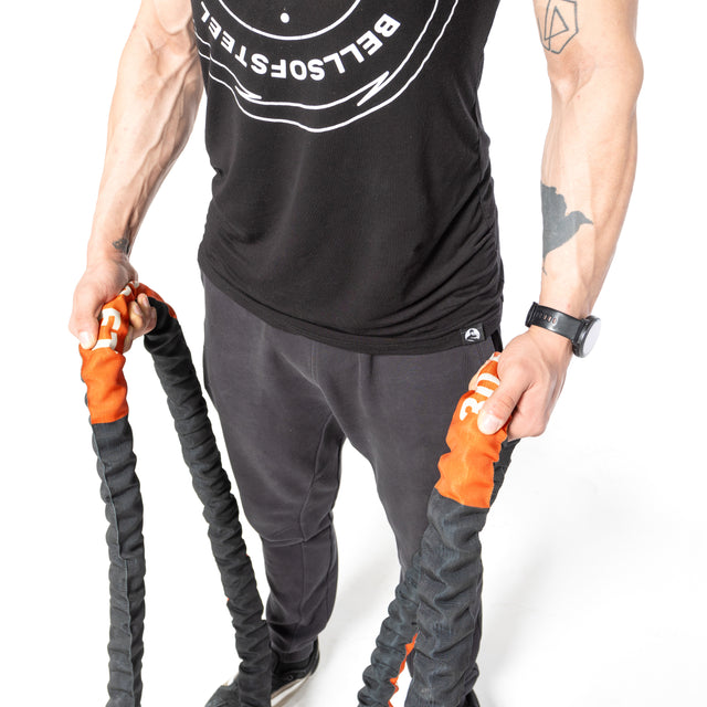 A person in a black tank top and pants grips Bells of Steel battle ropes, ready for an intense workout. Tattoos adorn their arms, complementing the watch on their left wrist as they prepare to engage with fitness equipment.