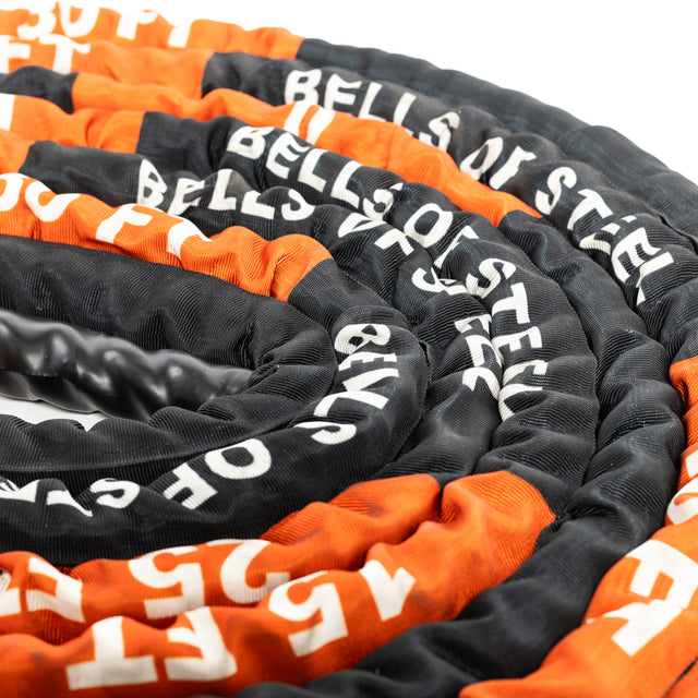 Close-up of coiled black and orange Battle Rope from Bells of Steel, printed with white letters. This essential fitness gear is arranged in a spiral, ideal for an intense workout.