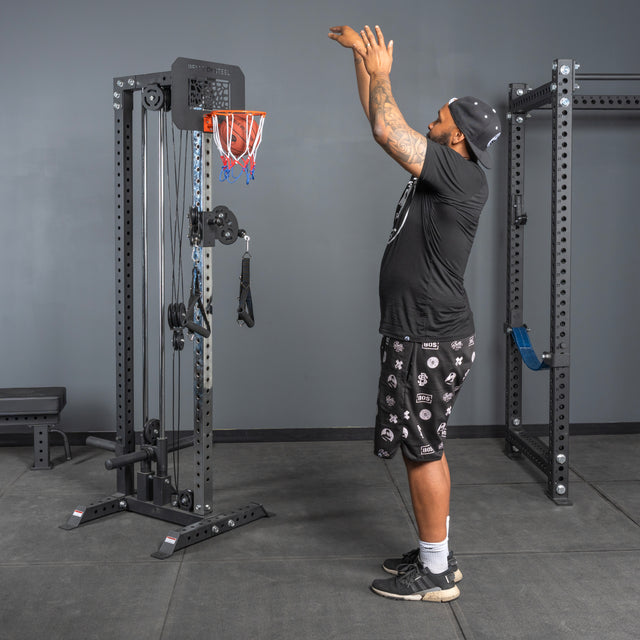 A man in athletic wear lines up a shot at the Bells of Steel Rack Attached Basketball Hoop w/ Ball, mounted on a gym cable machine in a fitness facility.