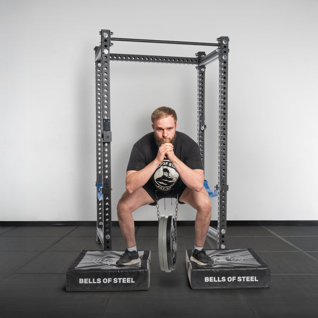 A man in athletic wear uses the Bells of Steel Dip Belt to perform a belt squat with a weight plate between two raised platforms in a gym, showcasing advanced bodyweight training techniques.