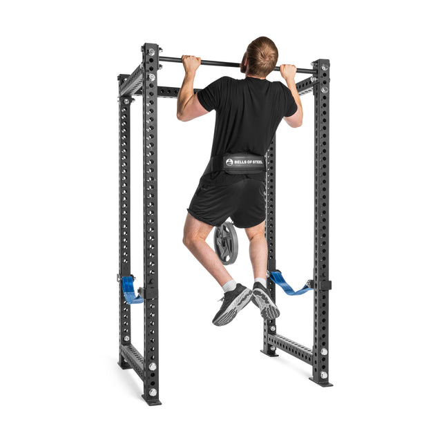 A man does a pull-up on a Bells of Steel power rack, wearing athletic gear and sneakers, with a weight plate attached to his waist using the Bells of Steel Dip Belt for advanced bodyweight training.