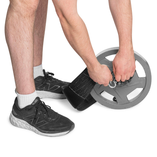 A person in black sneakers and white socks attaches a Bells of Steel Dip Belt to a gray weight plate; only their lower legs and hands are visible against a white background.