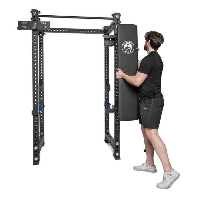 A man in sportswear attaches the Bells of Steel Wall or Rack Mounted Bench Hanger to a black metal power rack designed for strength training and gym storage, set against a plain white background.