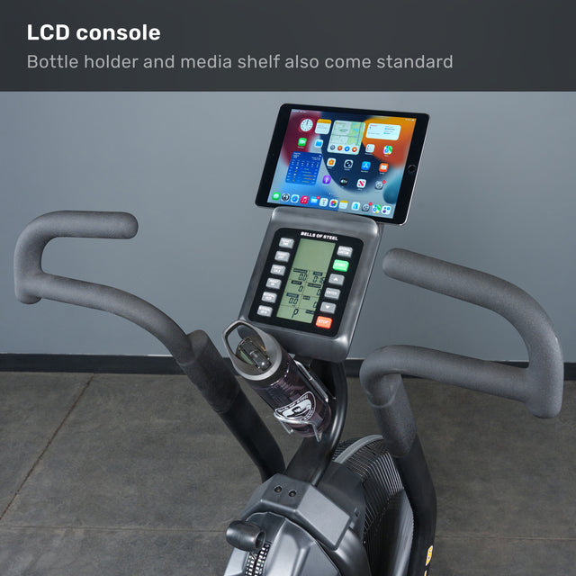 Close-up of the Bells of Steel Blitz Max Air Bike shows its LCD console with a media shelf for tablets and a bottle holder. The display presents workout stats, and text highlights features like the adjustable seat—ideal for any fitness studio.