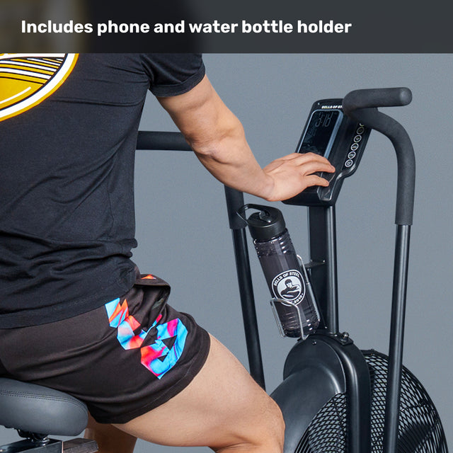 A person in a black shirt and colorful shorts uses the Bells of Steel Blitz Air Bike, pressing buttons on its display. A water bottle sits in the built-in holder. Text above reads, "Includes phone and water bottle holder.