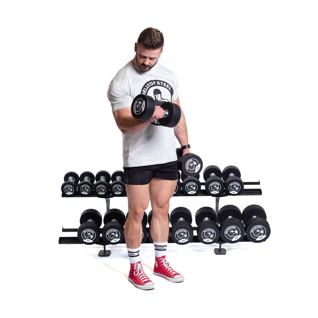 A man in a white t-shirt, black shorts, and red sneakers lifts a dumbbell with one arm, standing before the Bells of Steel Commercial Interchangeable Weight Rack filled with various weights on a white background.