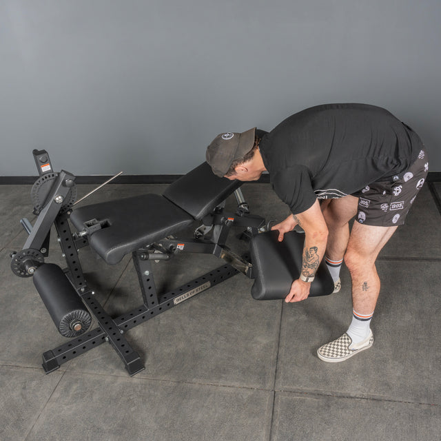 A person in casual clothes adjusts the padded section of the Bells of Steel Legacy Leg Extension / Hamstring Curl Machine - Plate Loaded in a home gym with gray walls and flooring.