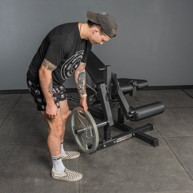 A person in black attire loads a weight plate onto the Bells of Steel Legacy Leg Extension / Hamstring Curl Machine - Plate Loaded in a gym with gray walls and floor.