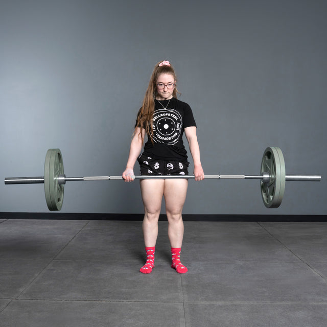 A woman in glasses, a black t-shirt and shorts, and red polka dot socks stands on a gray gym floor holding a Bells of Steel Deadlift Bar loaded with weights.