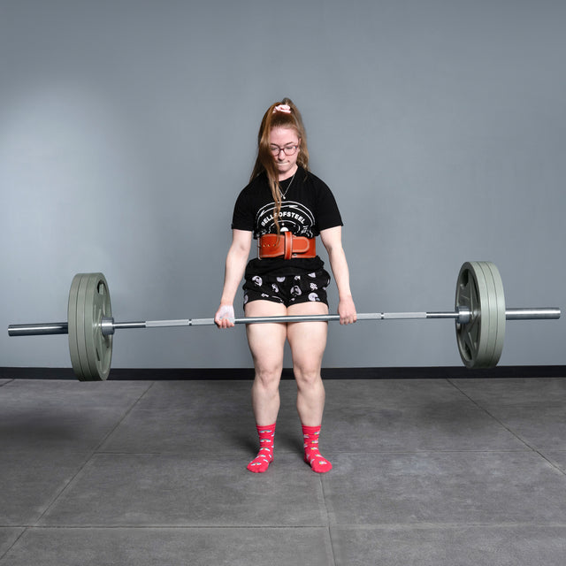 A person with long hair, glasses, and pink socks is powerlifting in a gym using the Bells of Steel Deadlift Bar, standing on a grey floor against a plain grey wall.