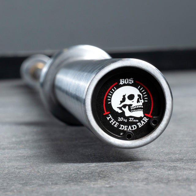 Close-up of a Bells of Steel Deadlift Bar end on the floor, featuring a skull graphic label with "BOS" and "20 kg 27 mm The Dead Bar," designed for powerlifting and inspired by deadlift bar standards.