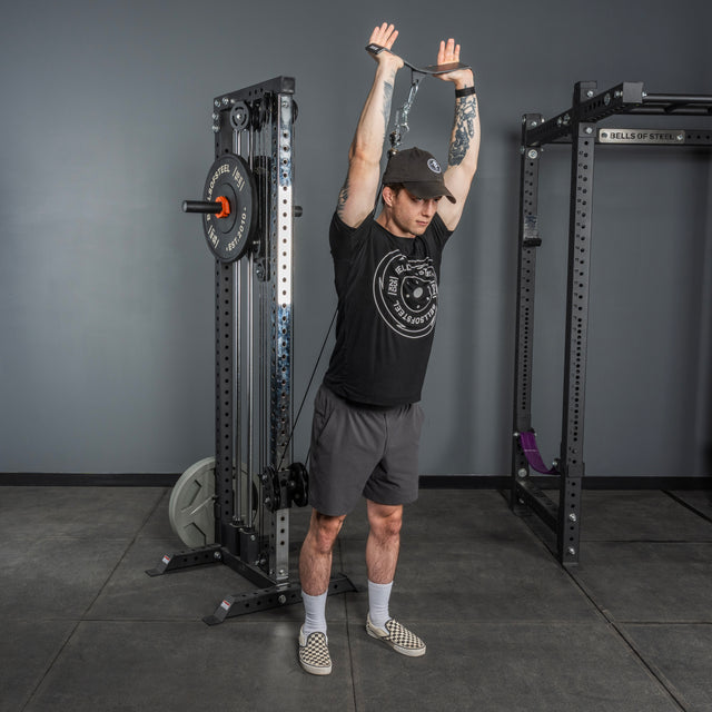 A man in a black cap, black t-shirt, and gray shorts uses the Bells of Steel Flat-Iron Tricep Press Cable Attachment to perform a standing overhead tricep extension in the gym, gripping the handle with both hands for optimal tricep engagement.