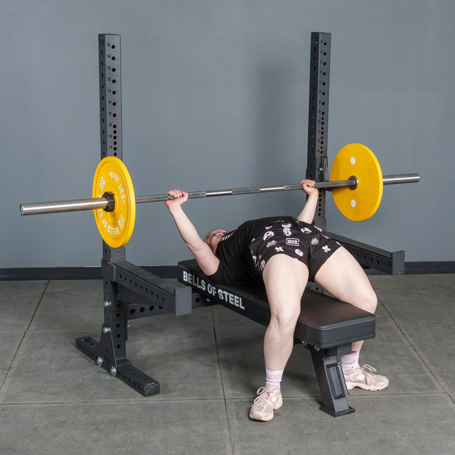 Wearing athletic clothing, a person bench presses a barbell with yellow plates on the Bells of Steel Hydra Two Post Converter Bench in a gym with gray walls and floor.