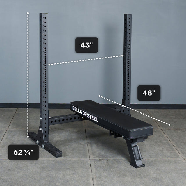 The Bells of Steel Hydra Two Post Converter Bench, a black bench with attached squat rack, is displayed on a concrete floor. “Bells of Steel” is printed on its side. Dimensions: 62¼" tall, 43" wide, and 48" deep.