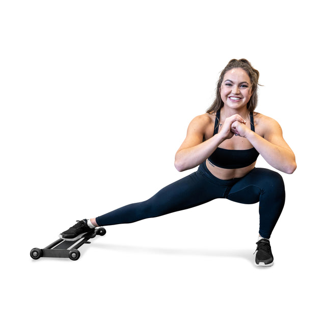 A woman in workout gear uses the Bells of Steel Glute Ham Slider, sliding her left foot on the wheeled platform while smiling and clasping her hands in front of her chest against a white background.