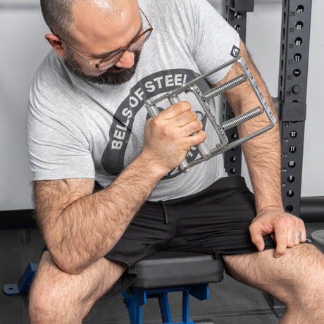 A bearded man in glasses, a gray "Bells of Steel" t-shirt, and black shorts uses the Bells of Steel Adjustable Forearm Heavy Trainer on a bench. Gym equipment is in the background, supporting his progressive overload routine.