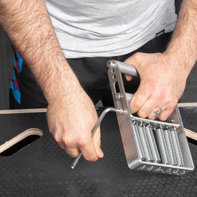 A person uses an Allen wrench to adjust the Bells of Steel Adjustable Forearm Heavy Trainer with multiple springs, placed on a black textured surface.