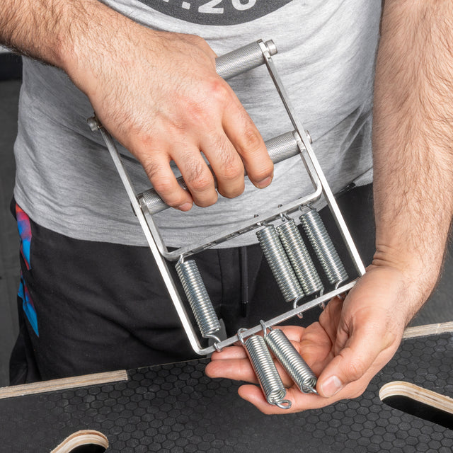 A person holds a Bells of Steel Adjustable Forearm Heavy Trainer grip tool with metal springs—one spring detached—while standing over a black textured surface.