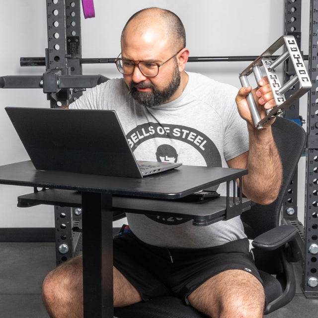 A man in a "Bells of Steel" t-shirt sits at his desk in a gym, working on his laptop while using the Bells of Steel Adjustable Forearm Heavy Trainer grip tool to focus on progressive overload and boost his grip strength.
