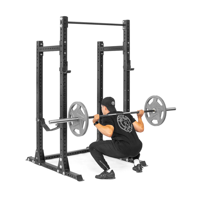 A person in black workout clothes and a cap performs a barbell squat inside the Bells of Steel Hydra Half Rack - Prebuilt (3" x 3", ⅝" Holes), with weight plates on the bar, demonstrating this durable rack’s suitability for any home gym.