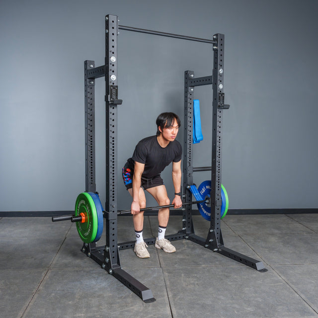 Wearing athletic gear, a person readies to lift a barbell with colorful plates inside the Bells of Steel Hydra Half Rack Builder (3" x 3", ⅝" Holes), ideal for home gyms, set in a room with gray walls and rubber flooring.