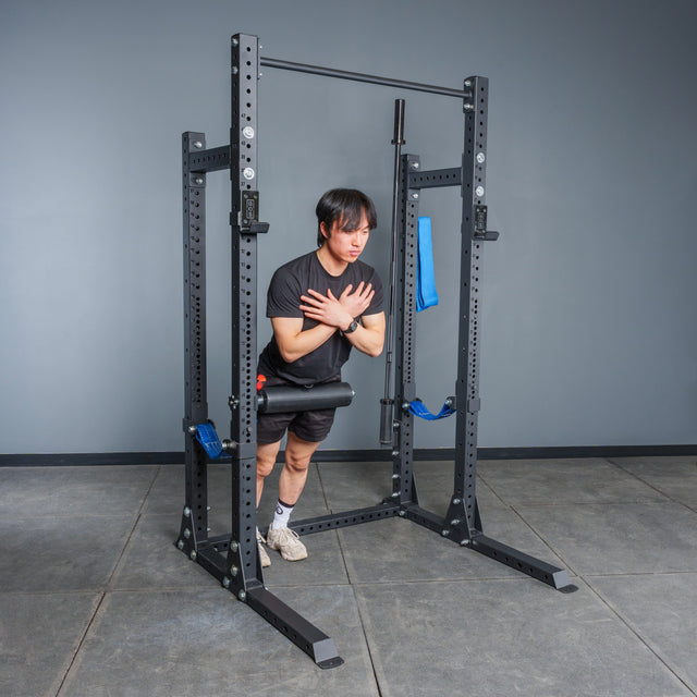 Using the Bells of Steel Hydra Half Rack Builder (3" x 3", ⅝" Holes), a person performs a Nordic hamstring curl in a gym, anchoring their feet under padded supports and crossing their arms over their chest.