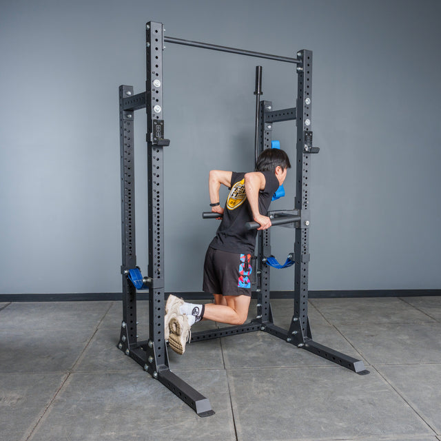 Wearing athletic clothing, a person performs a dip exercise on parallel bars attached to the Bells of Steel Hydra Half Rack Builder (3" x 3", ⅝" Holes) in a gym with gray walls and a tiled floor.