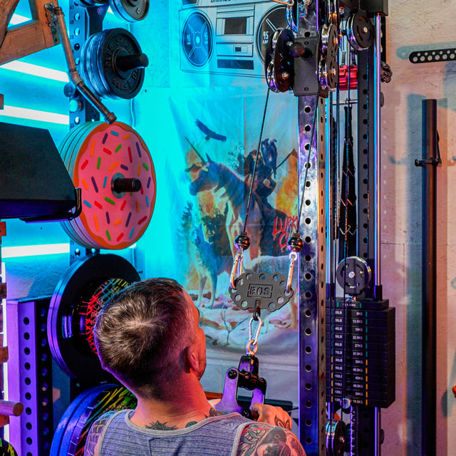 A man with short hair and a gray tank top works out on a home gym cable machine fitted with the Bells of Steel Kaizen Multi-Link, surrounded by neon lights, a donut-patterned weight plate, and a wall poster in a vibrant space.