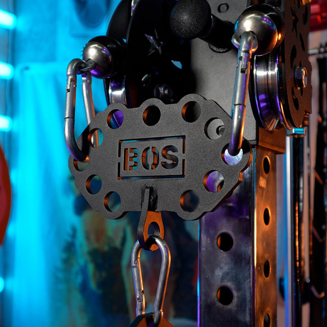 Close-up of the Bells of Steel Kaizen Multi-Link cable adapter, featuring "BOS" cut out on a metal plate, chrome details, and a carabiner. Blue and purple neon lights shine in the background—ideal for any home gym cable machine setup.