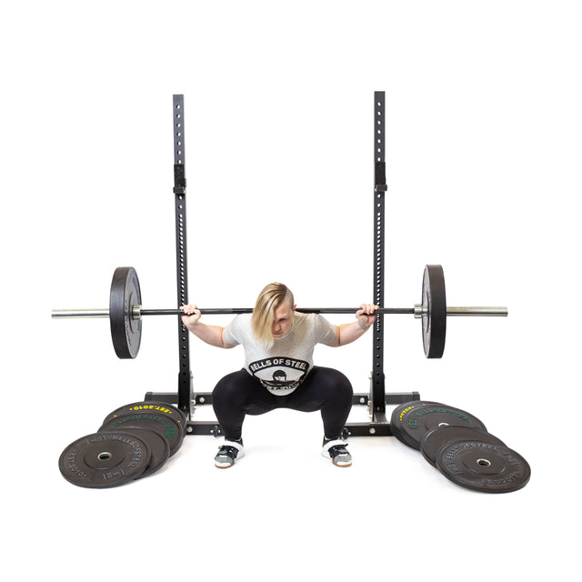 A person with blonde hair does a back squat using Bells of Steel Dead Bounce Conflict Bumper Plates on a barbell, supported by a squat rack with safety arms and surrounded by multiple weight plates for weightlifting.