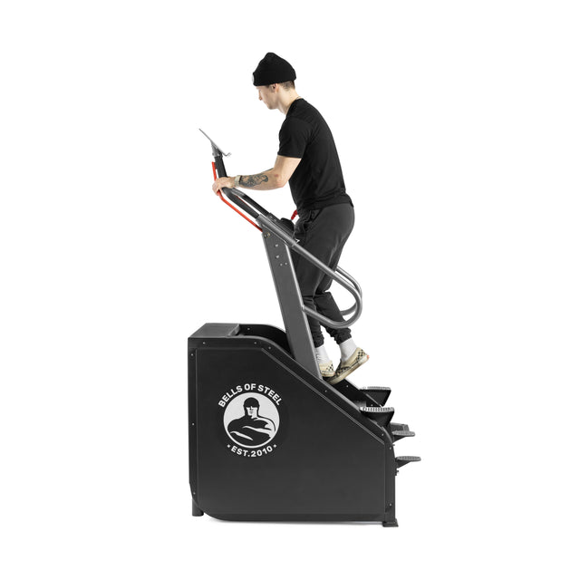 A person in a black shirt, pants, and beanie uses the Bells of Steel Sisyphean Stepper - Manual Stair Climber on a white background, demonstrating an effective cardio workout with adjustable resistance.