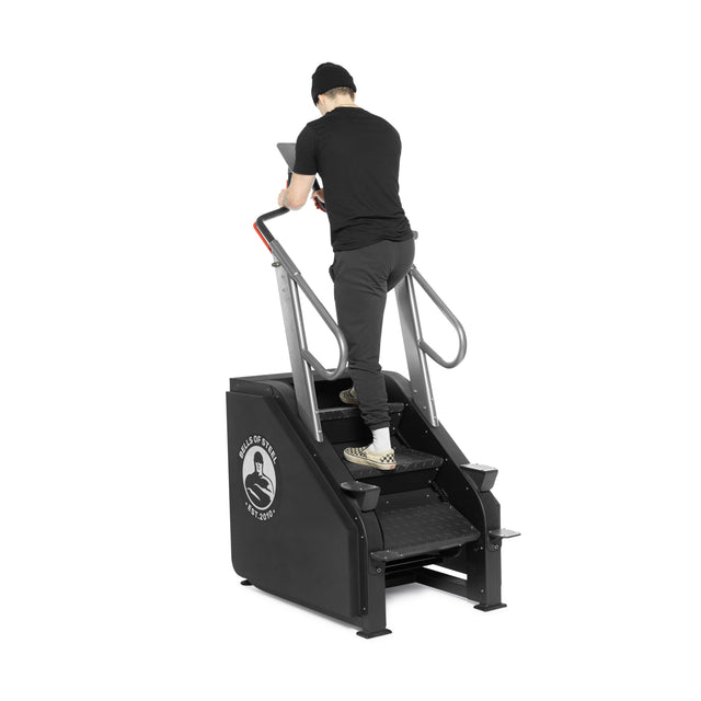 A person in black clothes and a beanie uses the Bells of Steel Sisyphean Stepper Manual Stair Climber with handrails and a logo visible on the side, in their home gym, facing away from the camera.