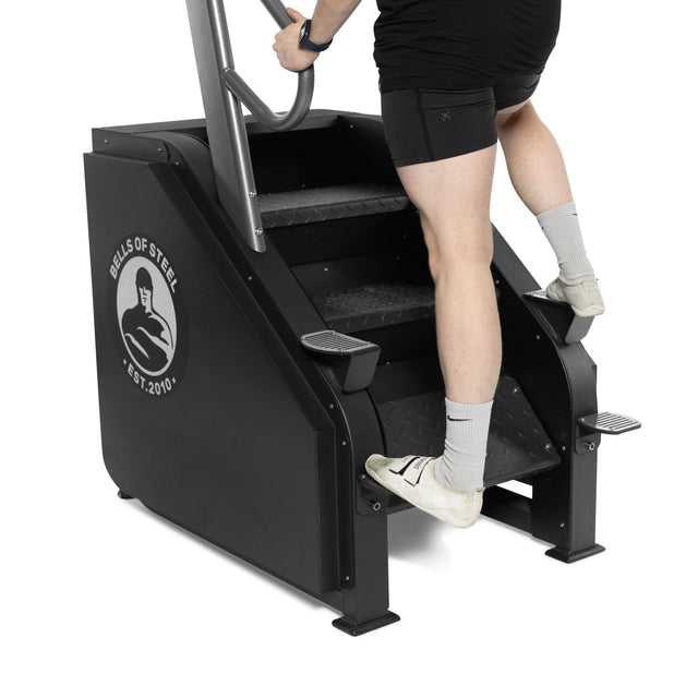 A person in athletic wear uses the Bells of Steel Sisyphean Stepper - Manual Stair Climber, gripping the handles and stepping on its black metal stairs. Only their lower body shows, highlighting this top cardio machine with adjustable resistance.