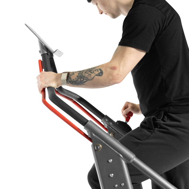 Dressed in black, a tattooed user grips the red handles of the Bells of Steel Sisyphean Stepper - Manual Stair Climber, smartwatch visible. Resistance levels are showcased on this top cardio machine against a white background.