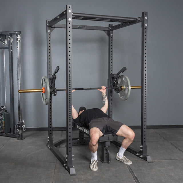 A person prepares to bench press on a bench inside a power rack equipped with the Bells of Steel Monolift Rack Attachment, gripping a barbell with weights in a gym setting.