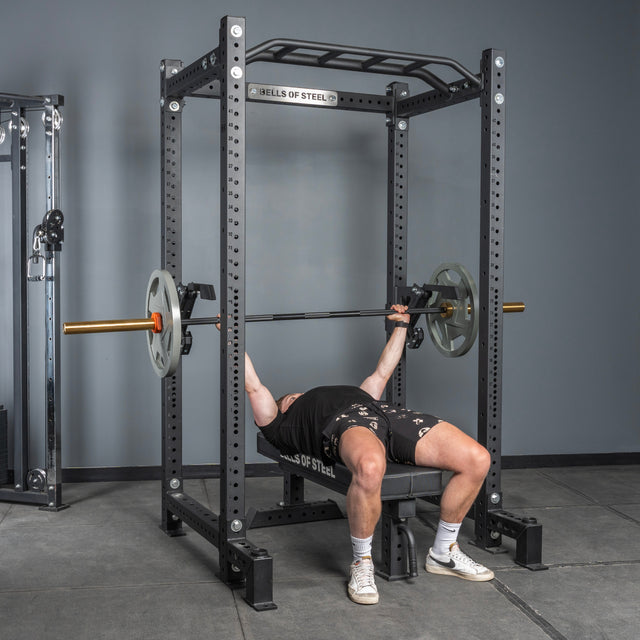 A person bench presses on a bench under a barbell, using the Bells of Steel Monolift Rack Attachment in a gym with gray walls and floor.