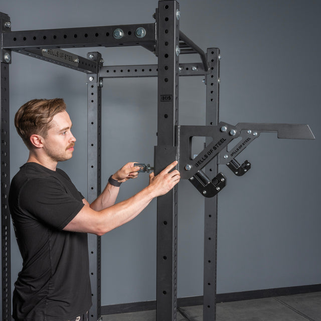 A man in a black shirt adjusts the Monolift Rack Attachment by Bells of Steel on a black power rack in a gym. The space-saving attachment features protective UHMW plastic for added durability.