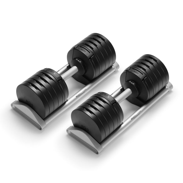 The Bells of Steel NÜOBELL-S Adjustable Dumbbells, featuring black plates and silver handles, rest side by side on sturdy stands against a white background—ideal for any home gym.
