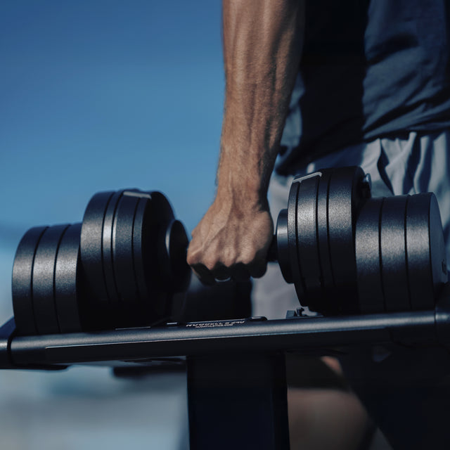 A close-up of a person’s hand gripping a Bells of Steel NÜOBELL-S Adjustable Dumbbell on a rack in a home gym, with the person in shorts and the background blurred.