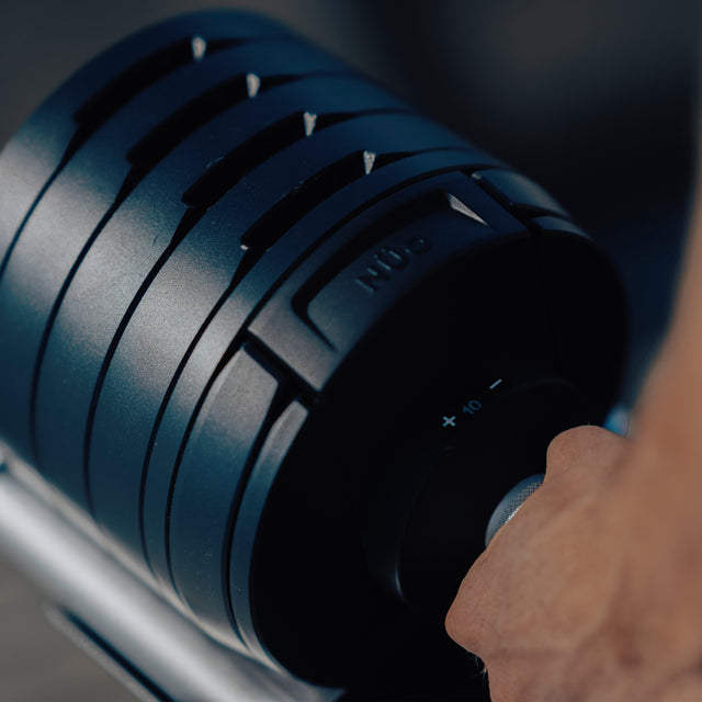A close-up shows a hand gripping the handle of a Bells of Steel NÜOBELL-S Adjustable Dumbbell, highlighting its precise weight settings and grooves—perfect for any home gym.