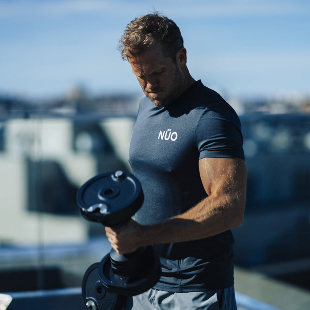 A short-haired man in a navy NUO t-shirt lifts Bells of Steel NÜOBELL-S Adjustable Dumbbells outdoors on a sunny day, focusing intently on his workout.