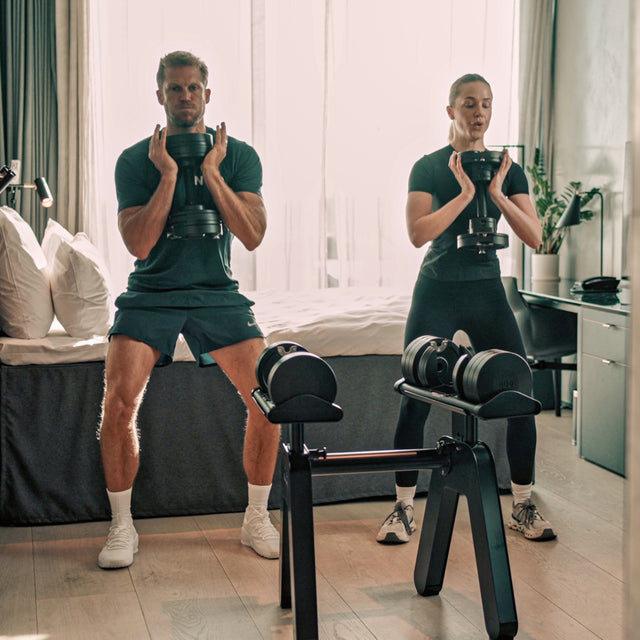 Two people exercise in a bedroom, performing squats and holding Bells of Steel NÜOBELL-S Adjustable Dumbbells. A set of these dumbbells rests on a stand in the foreground, as daylight filters through sheer curtains behind them.