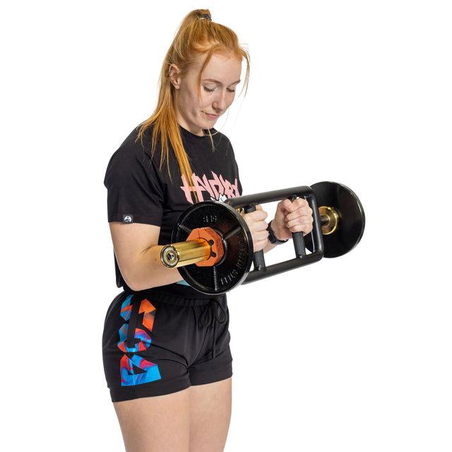 A woman with long red hair in a ponytail lifts a heavy barbell secured with Bells of Steel Magnetic Clamp Collars. She wears a black T-shirt with a pink-blue graphic and colorful shorts, standing against a white background.