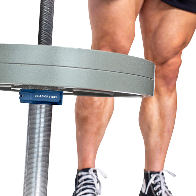 Close-up of a person's legs beside a loaded barbell secured with blue Bells of Steel Magnetic Clamp Collars. The person is wearing black shorts and black sneakers.