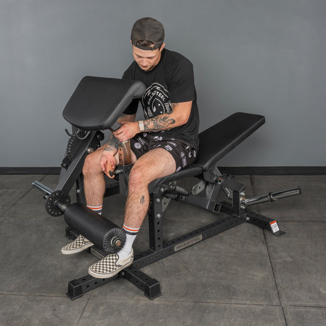 A man in a black shirt, shorts, and backward cap adjusts settings on a bench press in his home gym with gray walls and tiled floors. Nearby is the Bells of Steel Legacy Leg Extension / Hamstring Curl Machine - Plate Loaded.