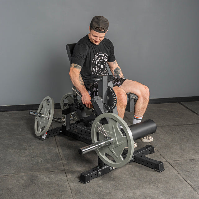 A man in a black t-shirt, shorts, and cap adjusts the Bells of Steel Legacy Leg Extension / Hamstring Curl Machine - Plate Loaded in a gym with large weight plates, gray flooring, and a plain wall background.