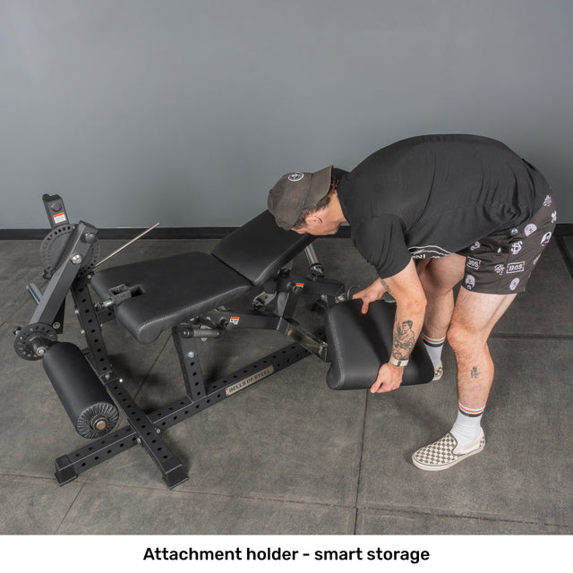 Wearing sportswear, a person adjusts the attachment holder on the Bells of Steel Legacy Leg Extension / Hamstring Curl Machine - Plate Loaded, showing smart storage for its accessories in a home gym.