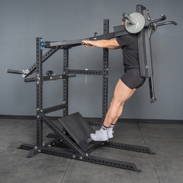 Wearing black athletic wear, someone performs a squat on the Bells of Steel Pandemonium Squat – 3-in-1 Pendulum Squat / Calf Press / Viking Press Machine, featuring padded shoulder supports and a slanted footplate in a gym with gray walls.